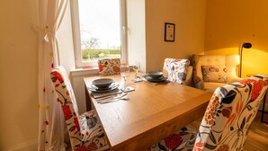 Uma área de refeições com mesa de madeira posta para dois, uma janela com cortinas e uma cadeira com padrão floral.
