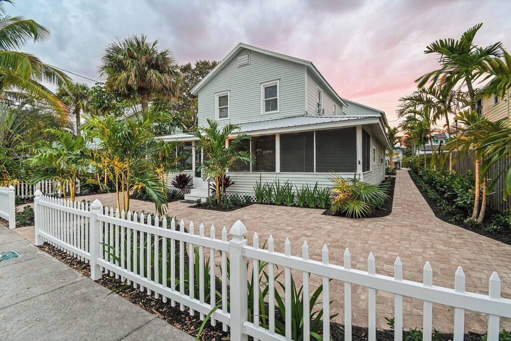 A two-story house with a covered porch, surrounded by a white picket fence and tropical plants.