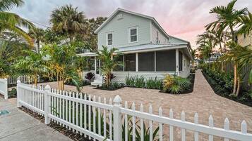 A two-story house with a covered porch, surrounded by a white picket fence and tropical plants.