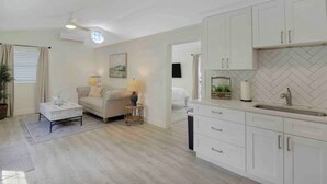 Un salon moderne comprenant un canapé, une table basse et une lampe, attenant à une cuisine avec des meubles blancs et un dosseret en motif chevrons.