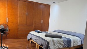 Un dormitorio con un armario de madera, una cama con colcha azul y blanca, y un ventilador de pie.