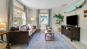 A living room with a sofa, coffee table, TV, and a surfboard on the wall. - Cottage 3 on Penn - Pool! (West Palm Beach)