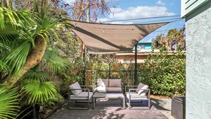 A patio with a sofa and chairs under a shade sail, surrounded by greenery and a building in the background. - Cottage 3 on Penn - Pool! (West Palm Beach)