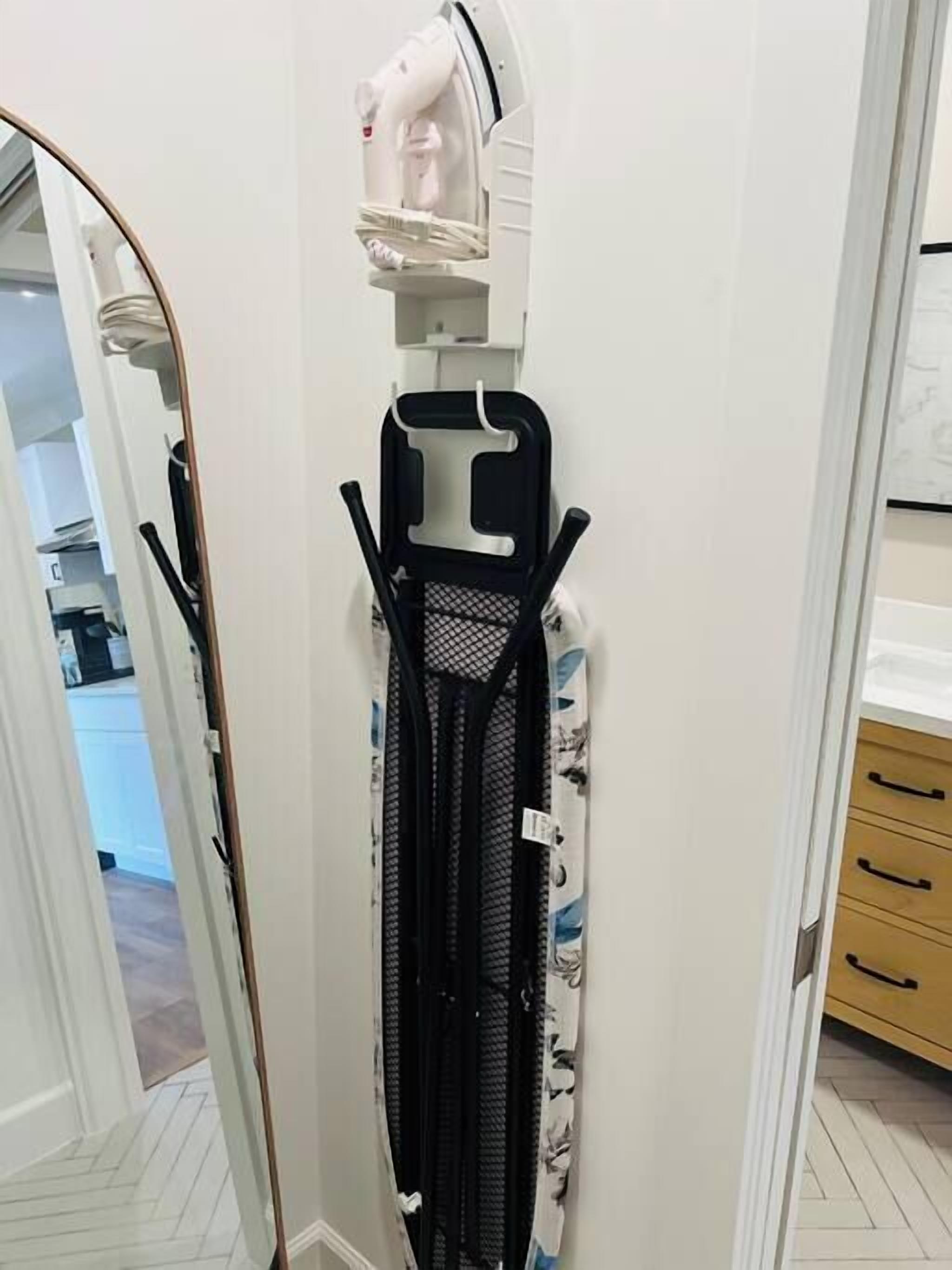 A compact iron rack with a mesh steamer attachment, a hanging iron, and a folded ironing board in a hallway.
