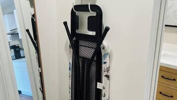 A compact iron rack with a mesh steamer attachment, a hanging iron, and a folded ironing board in a hallway.