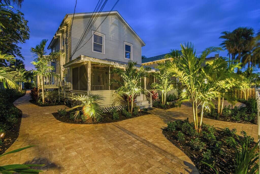 A two-story house with a covered porch, surrounded by tropical plants and a paved driveway.