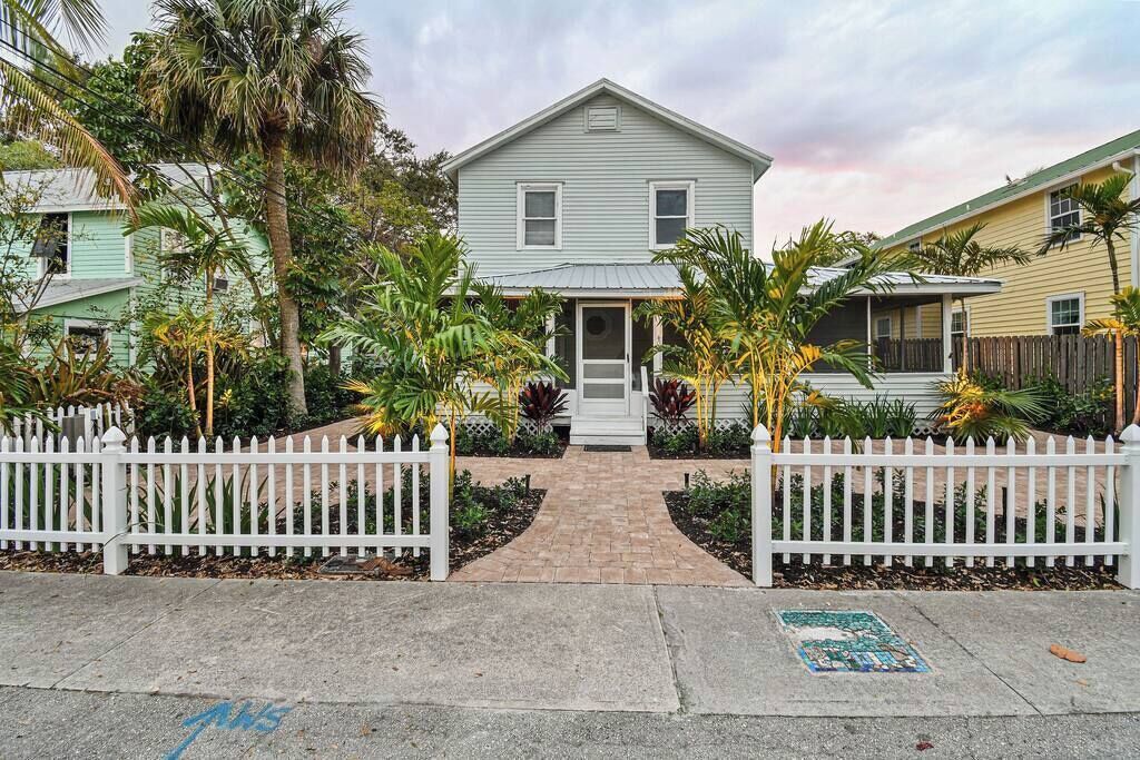 A two-story house with a front porch, surrounded by a white picket fence and tropical plants.