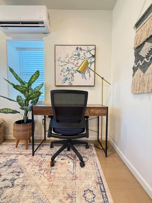 Un espace de travail comprenant un bureau, une chaise, un climatiseur, une plante en pot, une œuvre d’art et un tapis à motifs.