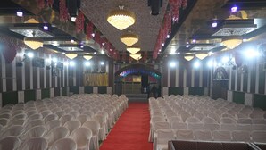 A hall with rows of white chairs on either side of a red carpet aisle, leading to a stage area.