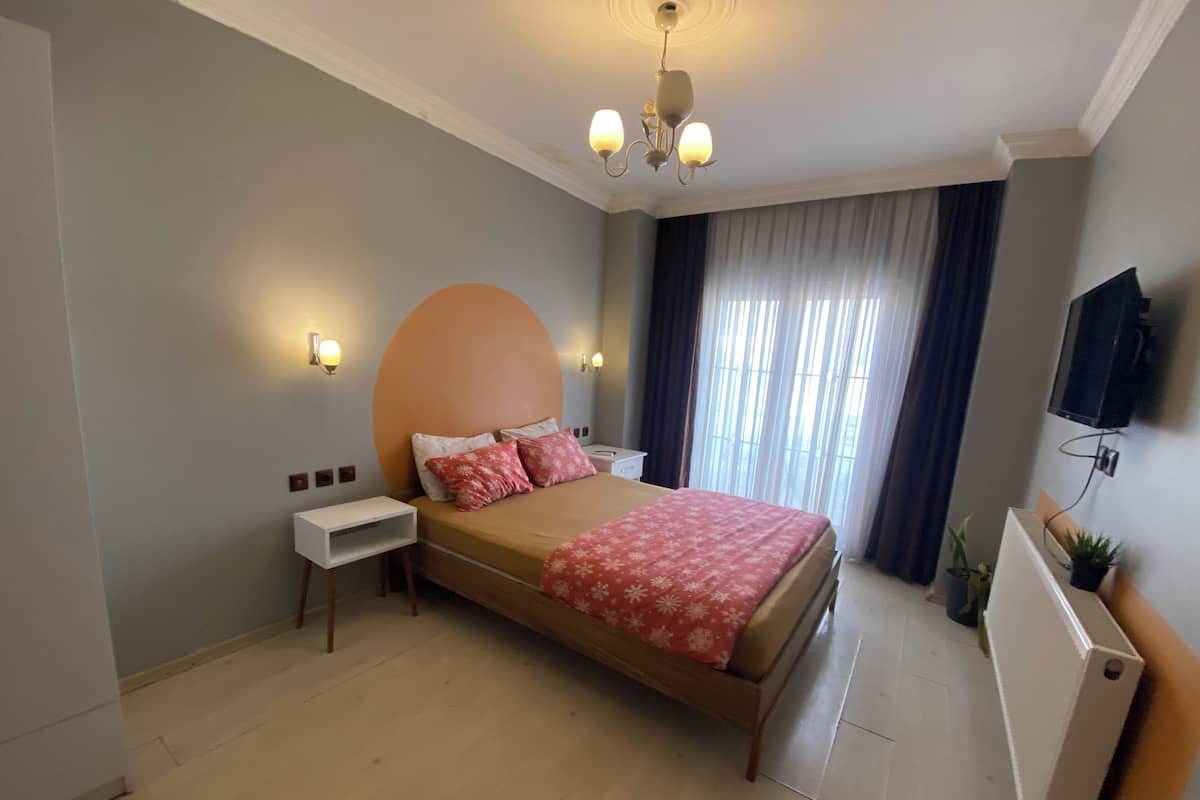 Un dormitorio con cama, mesitas de noche, televisor montado en la pared y ventana con cortinas.