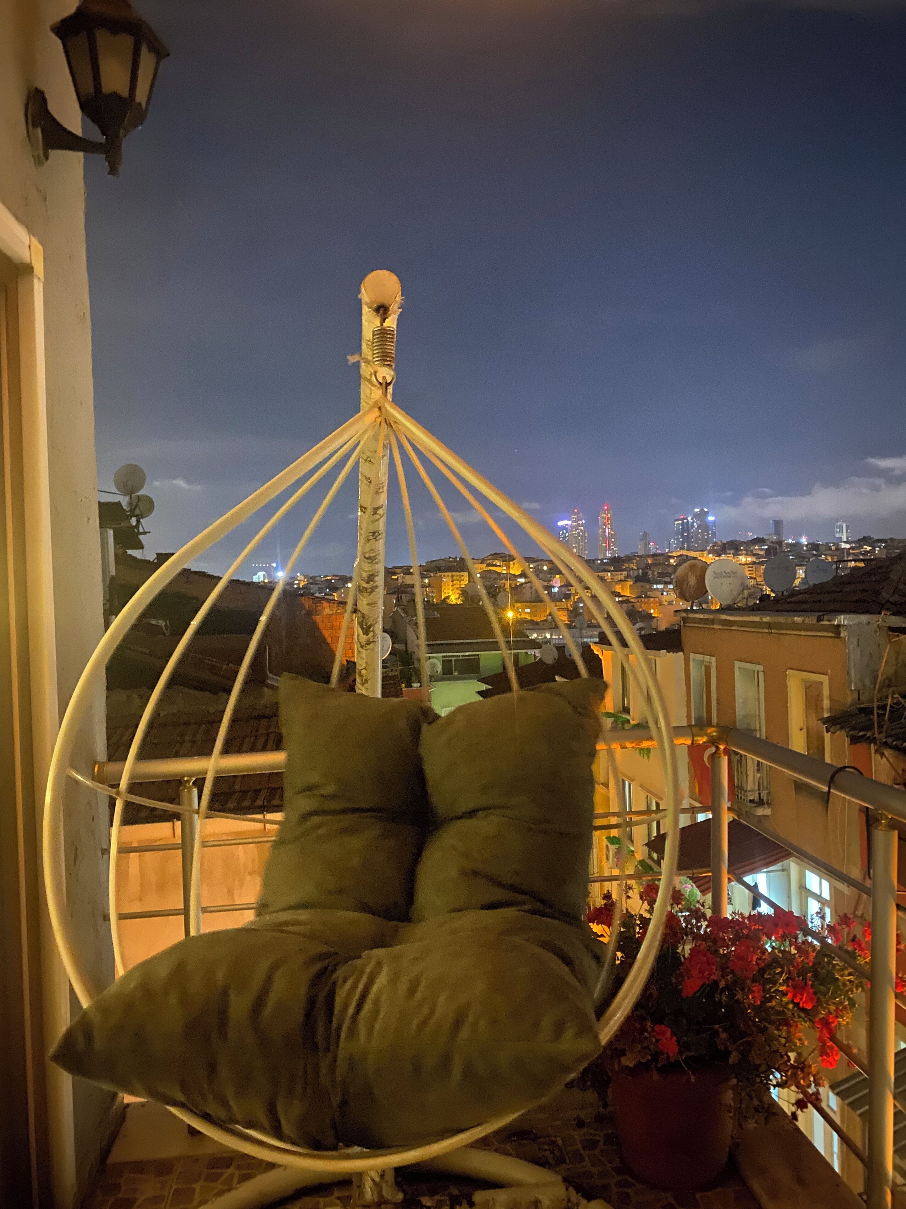 A hanging chair with cushions, a potted plant, and a cityscape view at dusk.