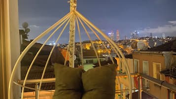 A hanging chair with cushions, a potted plant, and a cityscape view at dusk.