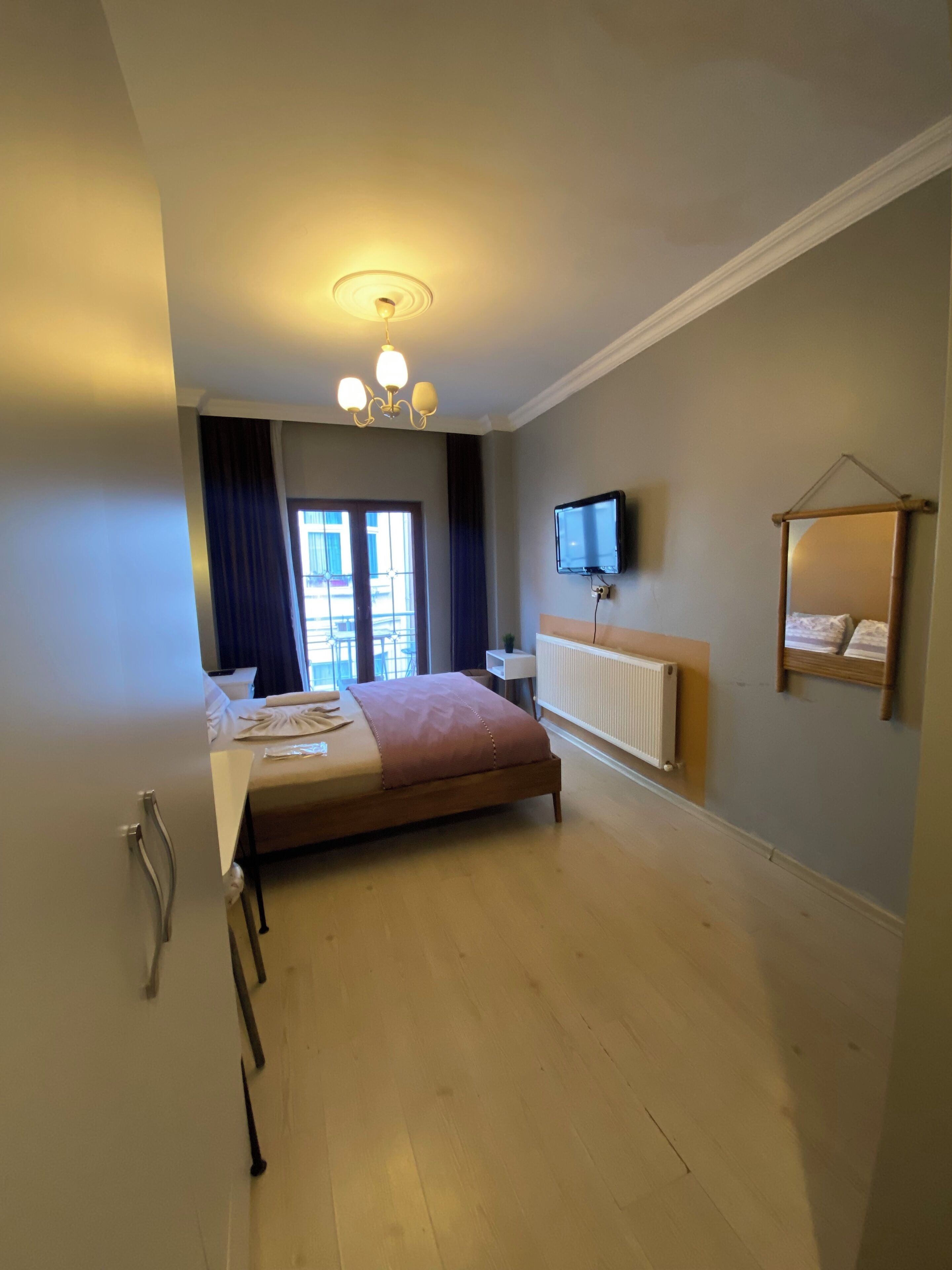 A hotel room with a bed, a TV, a radiator, a window with curtains, and a ceiling light fixture.