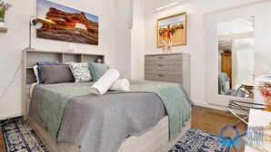 Um quarto com uma cama, mesa de cabeceira, abajur, pintura de uma paisagem, cômoda e espelho.