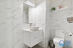 A bathroom with white marble tiles, a white sink with a gold faucet, a mirror, a toilet, and a small shelf with decorative items. - Mandurah Family Splash (Mandurah)