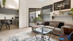 Uma sala de estar moderna com uma mesa de centro de vidro, um sofá e uma área de cozinha com armários cinzentos.