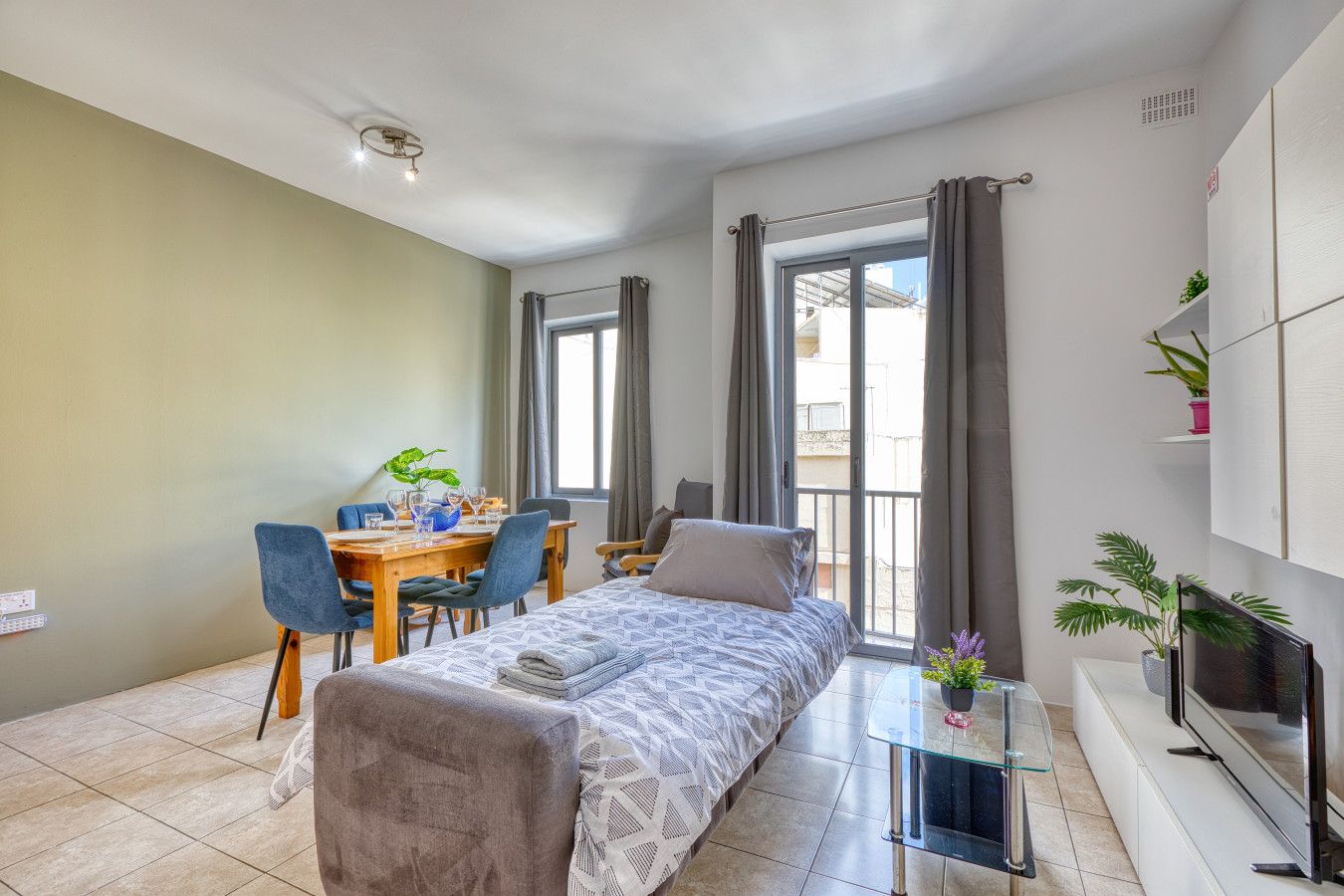 A modern bedroom with a bed, a dining table with chairs, a TV, and a balcony with curtains.