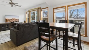 Een moderne woonkamer met een grijze bank, een houten eettafel en stoelen. Er is een plafondventilator, grote ramen en een televisie op een dressoir.