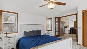 Een slaapkamer met een houten hoofdbord, een bed met een blauwe deken, een nachtkastje, een dressoir, een plafondventilator en uitzicht op de aangrenzende keuken.