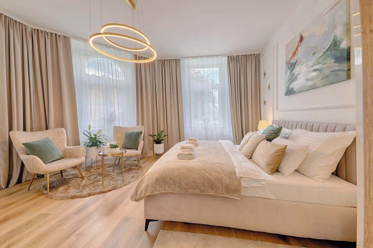 A modern bedroom with a large bed, two armchairs, a round coffee table, and a painting on the wall.