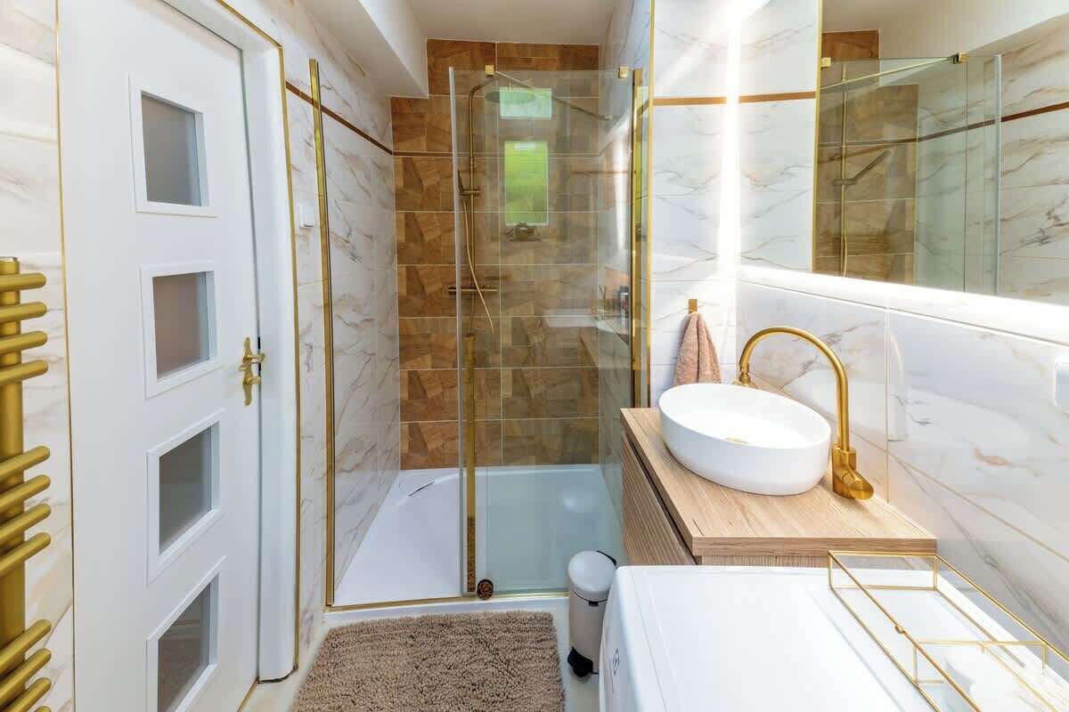 Ein modernes Badezimmer mit einer gläsernen Duschkabine, einem weißen Waschbecken mit goldfarbenen Armaturen und einem marmorbelagten Duschbereich.