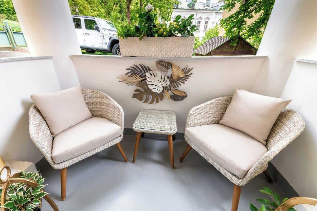 A balcony with two wicker chairs and a small table, a decorative wall with leaf patterns, and a white car parked outside.