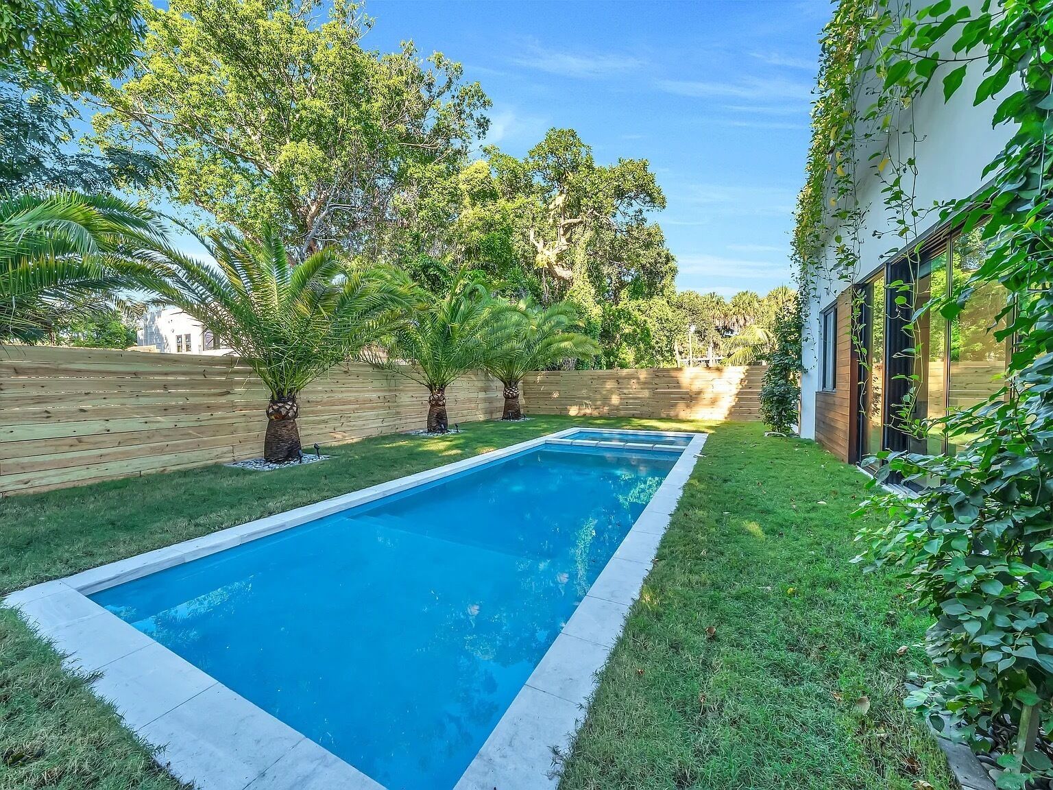 A rectangular swimming pool with a wooden deck surrounding it, set against a backdrop of lush greenery and a wooden fence.