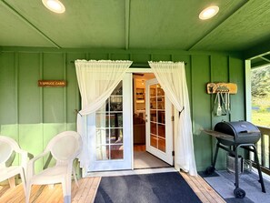 A covered porch with a grill, two chairs, a door leading to a kitchen, and a sign indicating '1 SPRUCE CABIN'. - ‘Spruce’ Riverfront Cabin near Olympic National Park (Beaver)