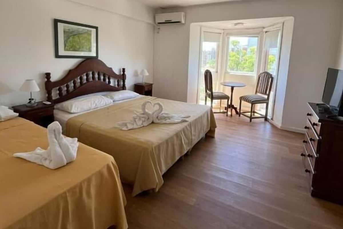 A hotel room with two beds, a wooden headboard, a desk, a chair, and a window with a view of greenery.