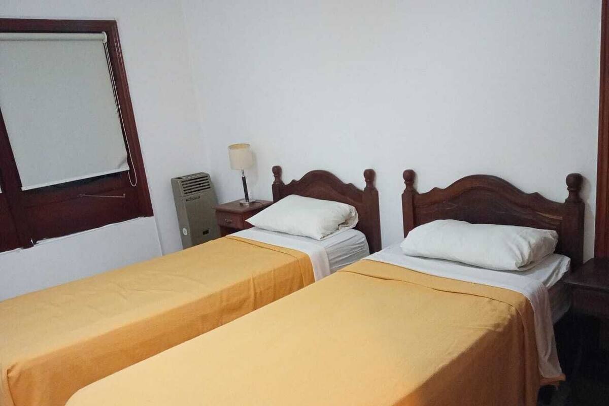 A room with two single beds, a bedside table with a lamp, a heater, and a window with a blind.