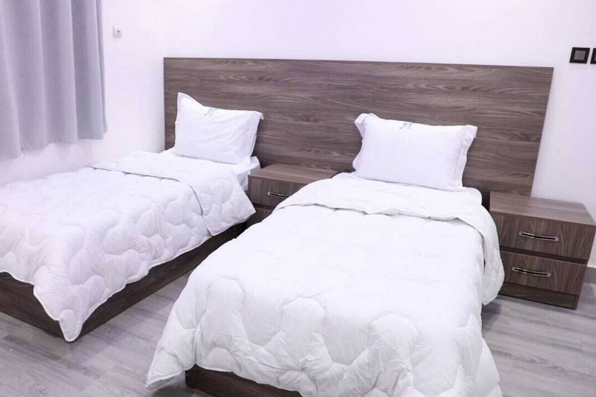Una habitación con dos camas individuales, cada una con cabecero de madera, ropa de cama blanca y una mesita de noche pequeña con cajones.