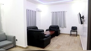 A modern living room with a grey sofa, two black armchairs, a wooden coffee table, and a TV mounted on the wall. - Hotel Tijiratt (Nouakchott)
