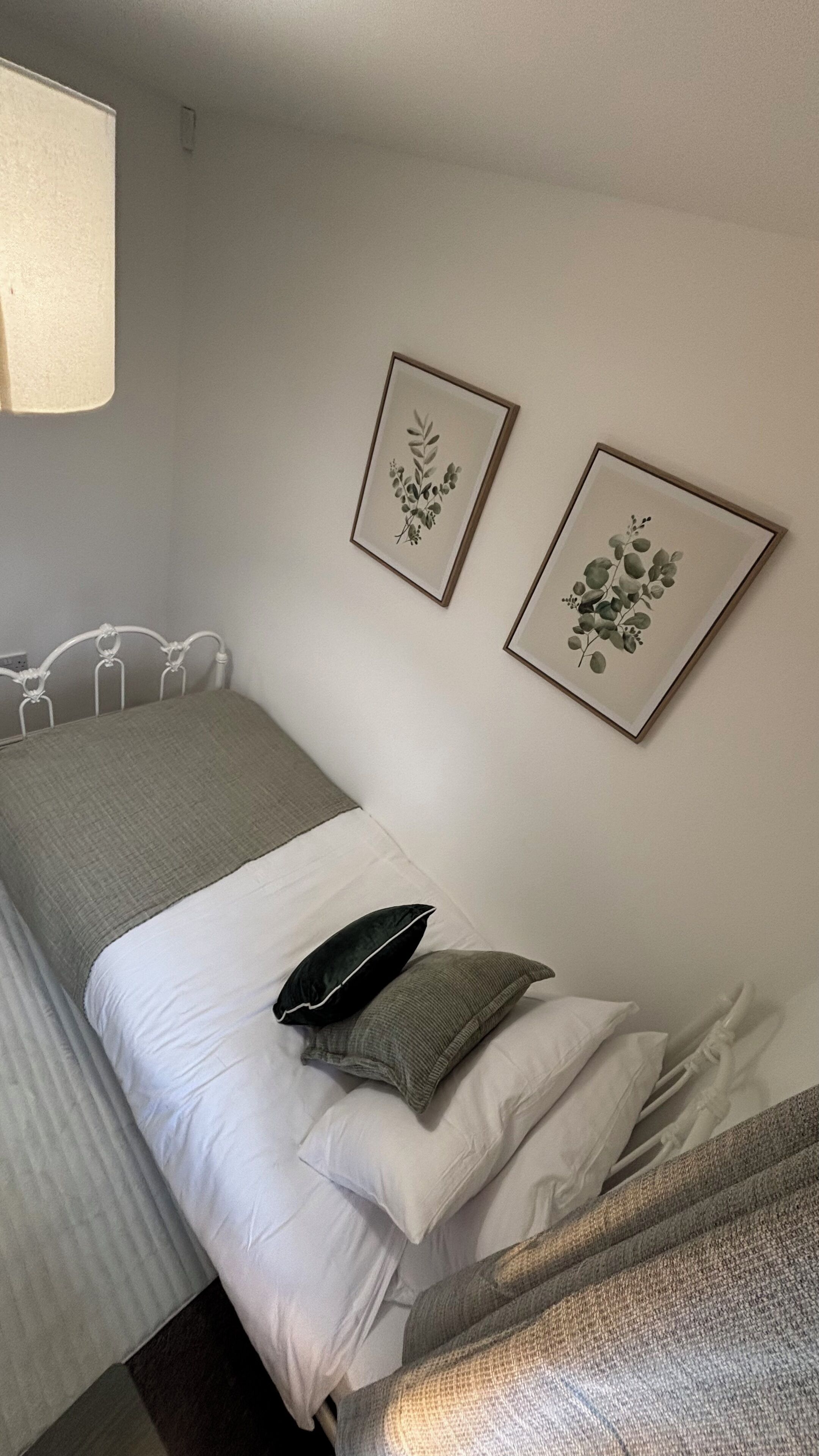 Una cama pequeña y ordenada con una colcha blanco y gris, dos almohadas y dos láminas botánicas enmarcadas en la pared.