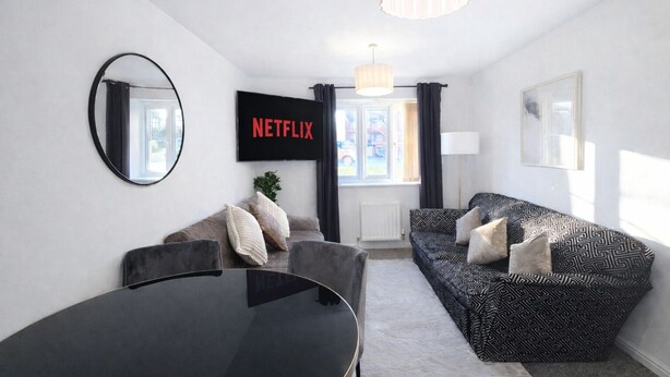 Een moderne woonkamer met een zwarte eettafel, twee banken, een grote spiegel, een televisiescherm waarop 'NETFLIX' staat, en een raam met gordijnen.
