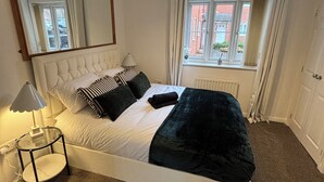 Een slaapkamer met een groot bed, een nachtkastje, een lamp, een raam met gordijnen en een spiegel aan de muur.