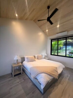 Ein modernes Schlafzimmer mit Bett, Nachttischen, Deckenventilator und einem Fenster mit Blick auf Grünflächen.