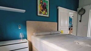 Een slaapkamer met een bed, nachtkastje, lamp, klok en een schilderij aan de muur.