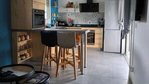 Een moderne keuken met een eethoek, met een inbouwoven, een magnetron en een koelkast. Er is een houten kast en een plank met keukengerei.