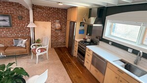 Eine kompakte Küche mit Backsteinwand, Holzboden, einem Essbereich mit Sofa und Stühlen sowie einem großen Fenster.
