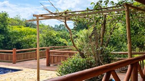 A wooden deck with a pergola structure, a pool, and lush greenery in the background.