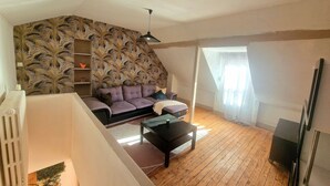 A loft-style living room with a sloped ceiling, a built-in bookshelf, a purple sofa, a coffee table, and a TV on a stand. - Cozy House Near the Castle & Lakeside Leisure Area (Cepoy)