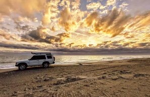 Un SUV blanco estacionado en una playa arenosa con un atardecer de fondo.