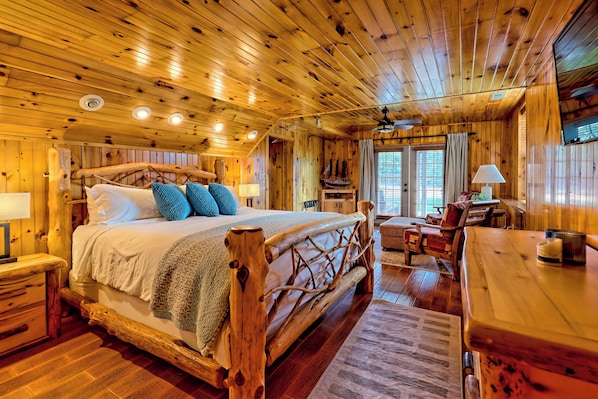 A wooden cabin interior with a large bed, a wooden desk, and a dining table.