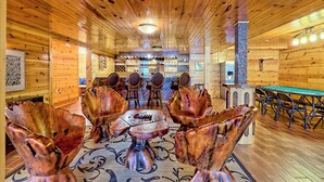 A wooden interior with a central table surrounded by unique copper chairs, a bar area, and a fireplace. - Lakefront Luxury w/ Dock + Fenced Yard (Blairsville)