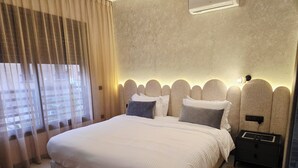 Un dormitorio moderno con una cama grande, cabecera con diseño texturizado, mesita de noche con toma de corriente y una ventana con cortinas traslúcidas.