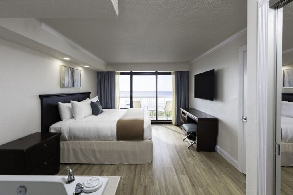 A hotel room with a bed, bedside table, TV, and a balcony with a view of the ocean.