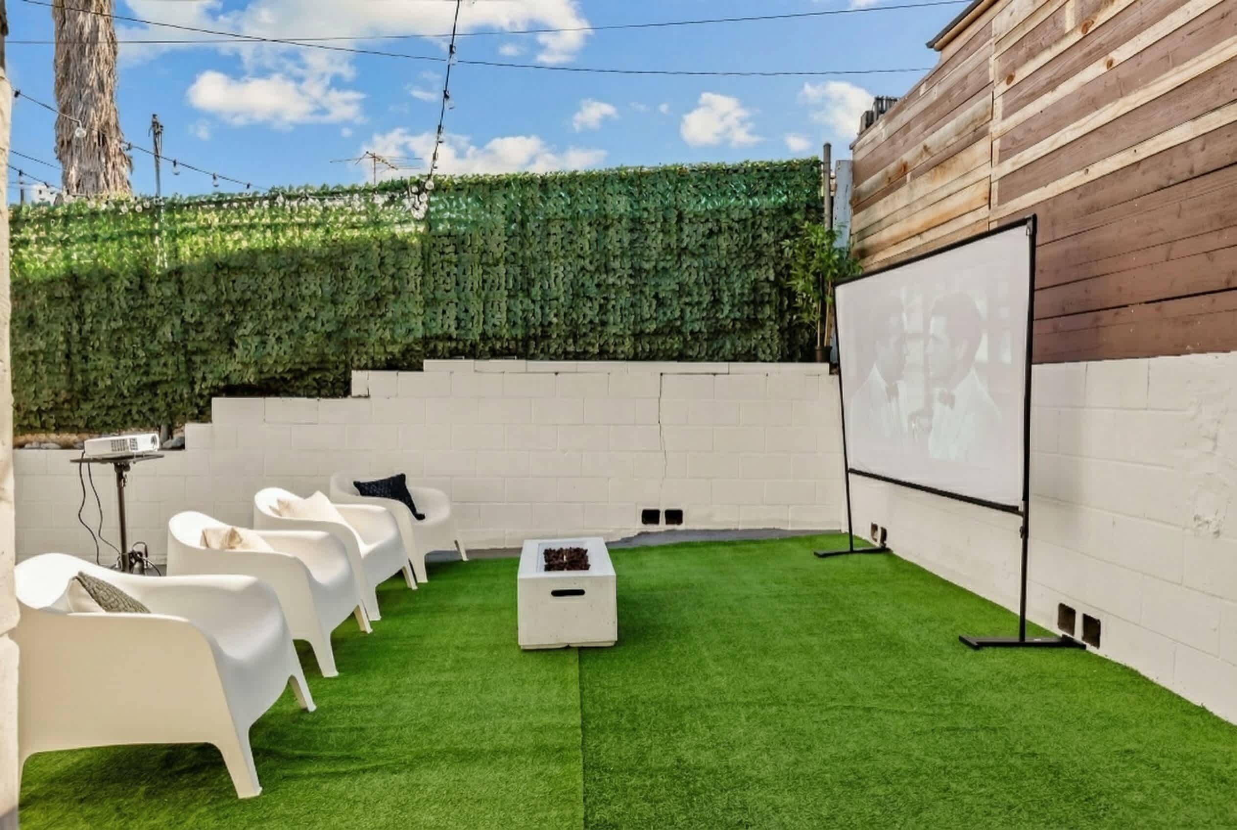 Un jardin avec une pelouse artificielle, des fauteuils de jardin blancs, un écran de projection et un mur végétal.