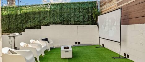 Un jardin avec une pelouse artificielle, des fauteuils de jardin blancs, un écran de projection et un mur végétal.
