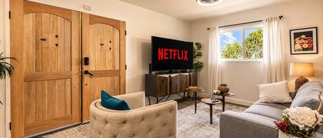 A living room with a television, a sofa, a chair, a coffee table, and a large window with curtains.