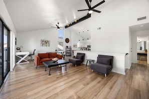 A modern living room with a wooden floor, a black ceiling fan, and a kitchen in the background. - Brilliant 3-bedroom townhome in charming April Sounds Community (Montgomery)
